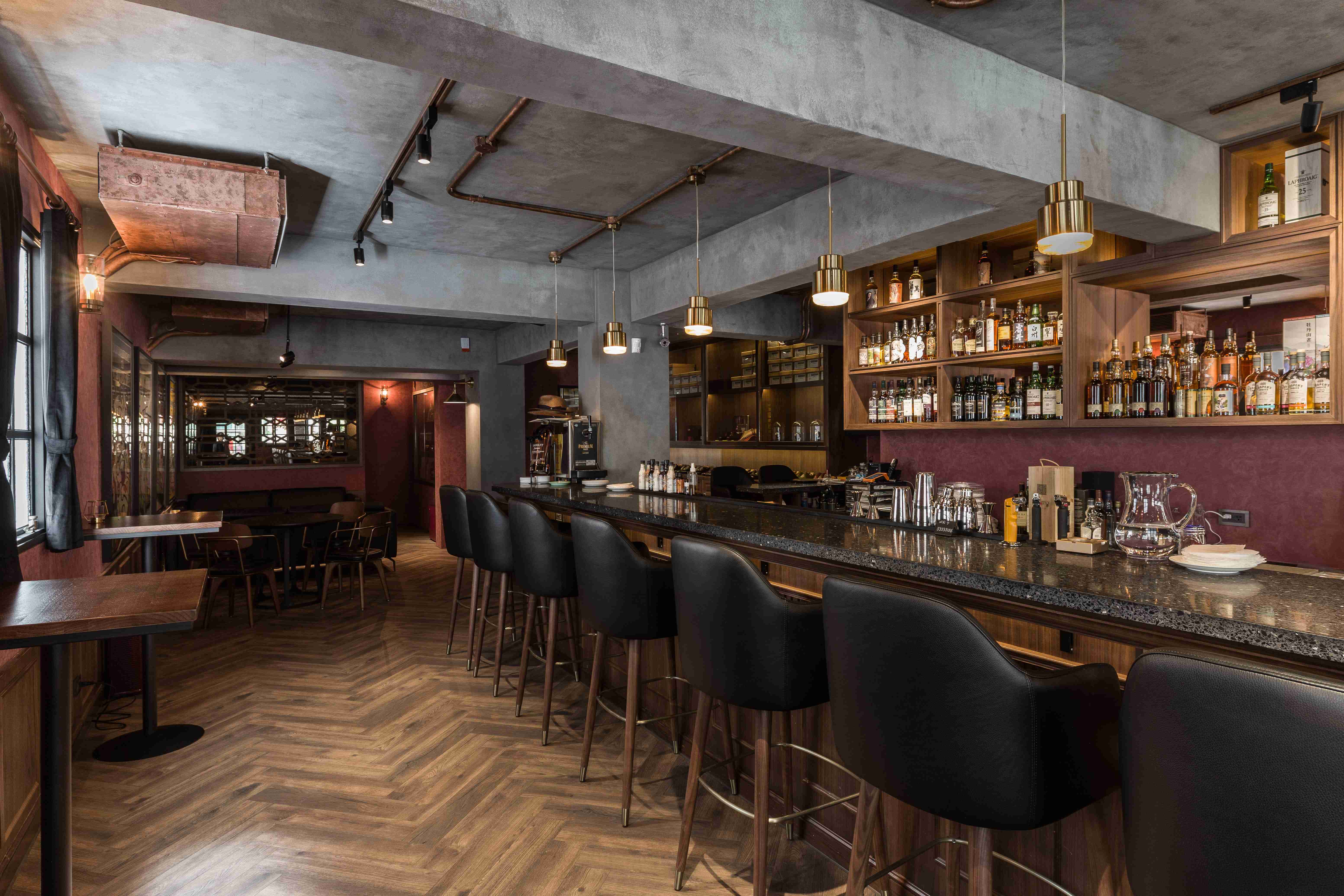 本案酒吧設計以長型大理石吧台為主軸，搭配黑色皮革高腳椅，營造舒適高級的品酒空間。紅銅色牆面與黑框窗戶帶出濃厚的復古氛圍，搭配工業風吊燈與牆面燈飾，增添溫暖神秘感。
